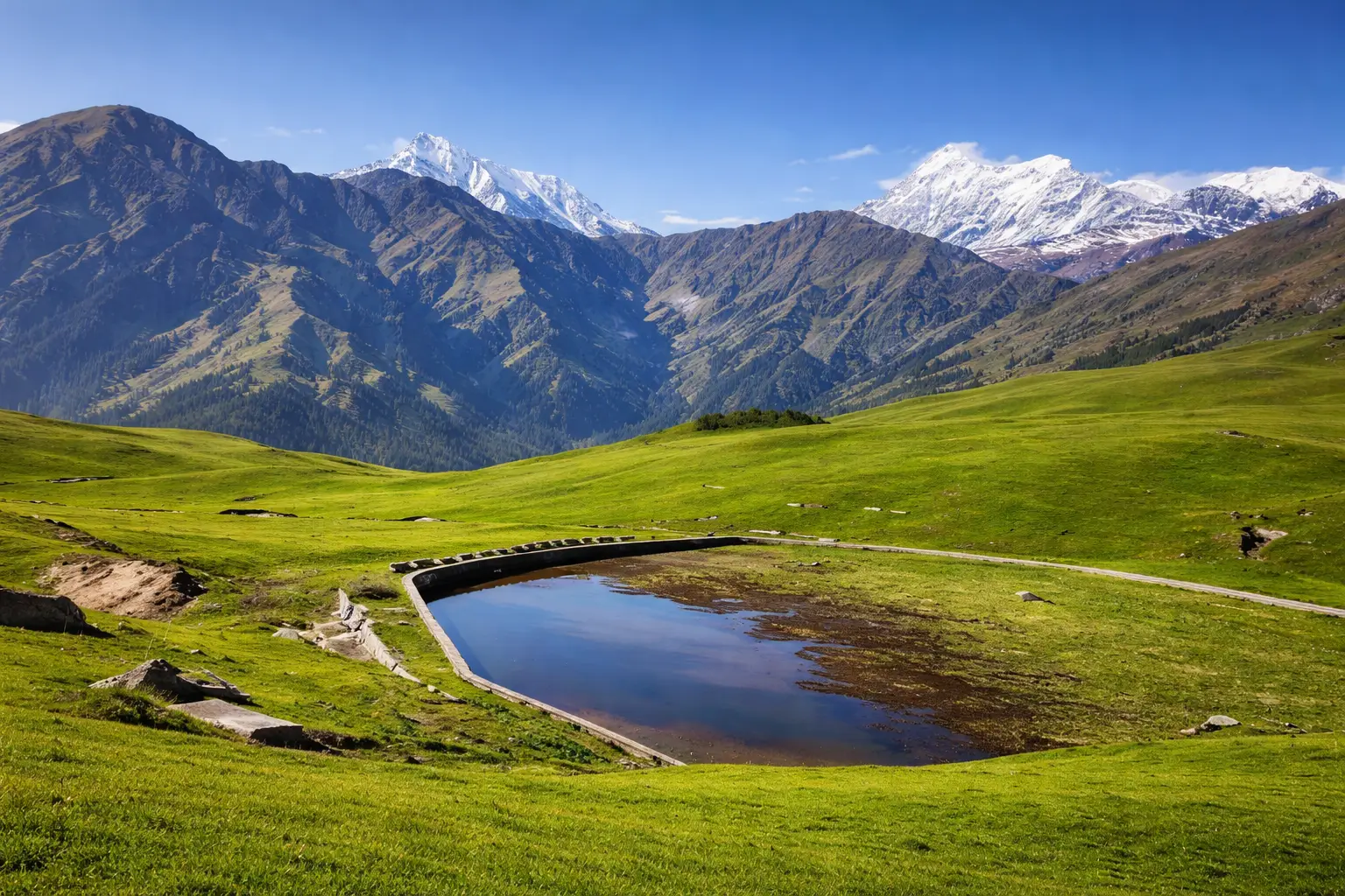 Ali Bedni Bugyal alpine meadow with green grassland, water pond and light snow covered Himalayan mountains under blue sky